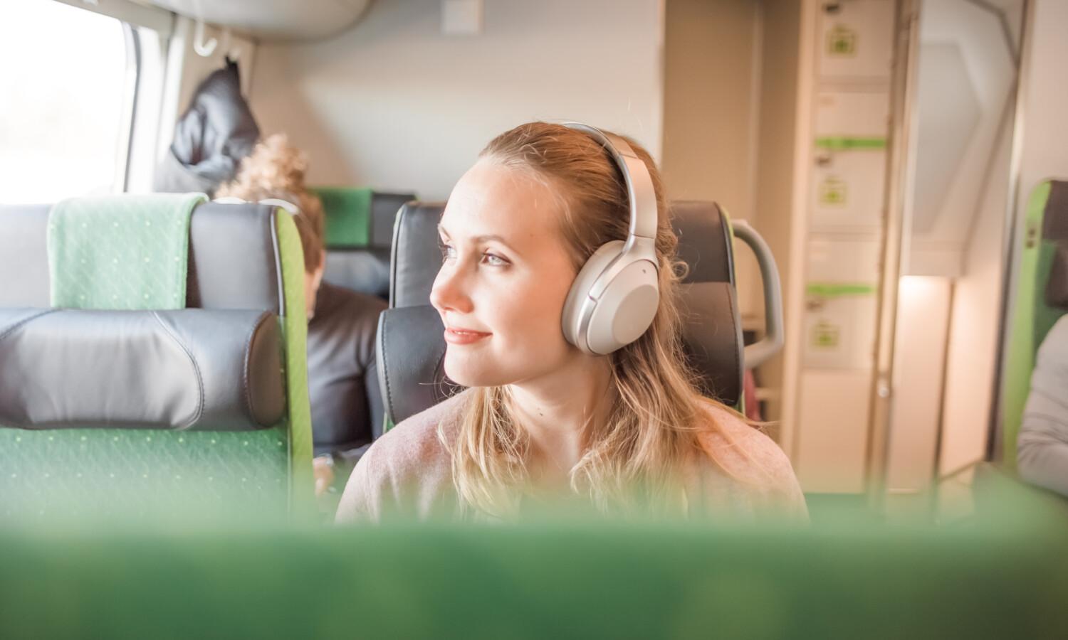 Nainen istuu junassa kuulokkeet korvillaan ja hymyilee